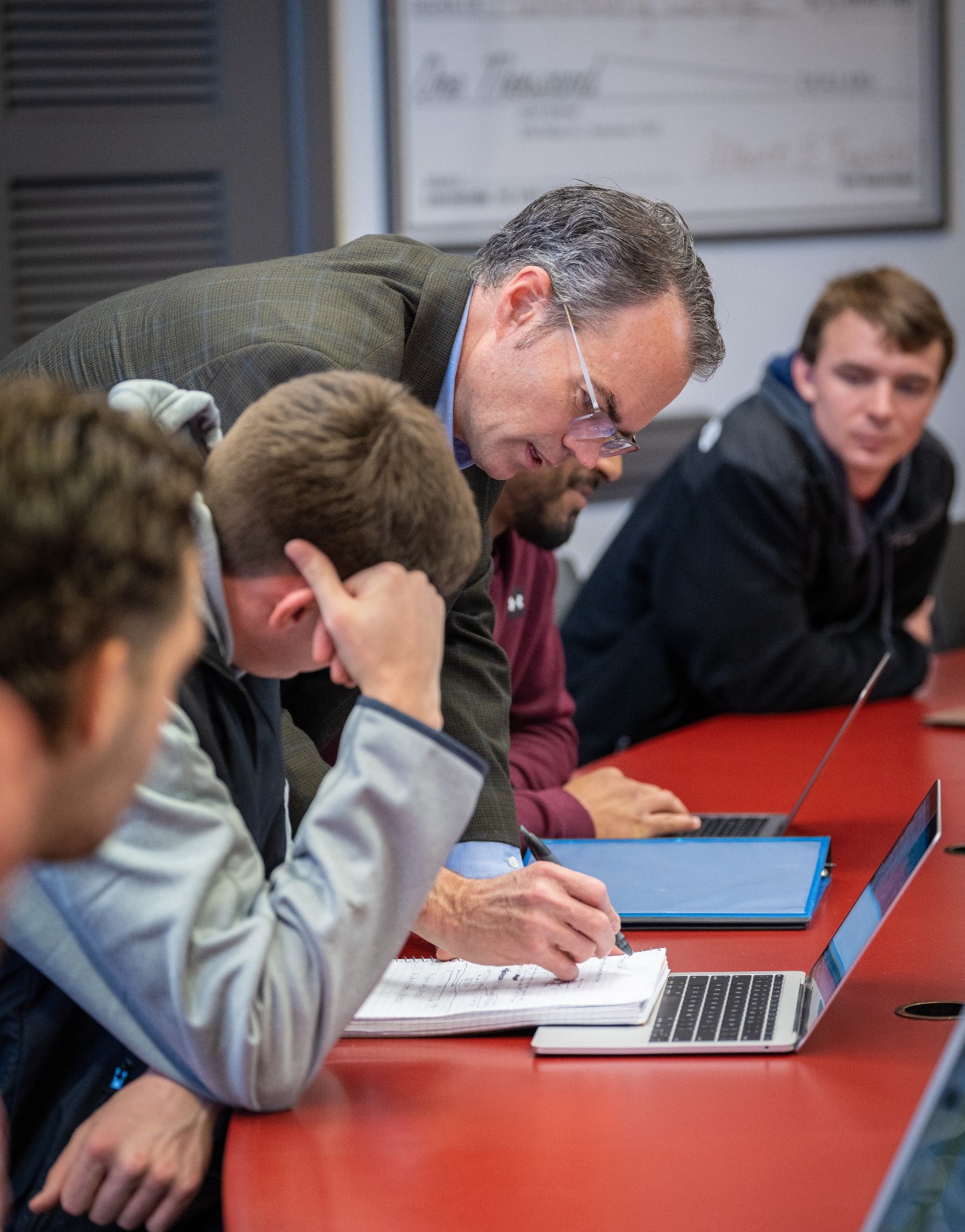 An instructor leans over the shoulder of a student and writes something on the paper in front of them while other students look on.